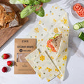 Beeswax wraps with a sandwich and vegetables on a white surface, next to a package of ZWS Beeswax Wraps.