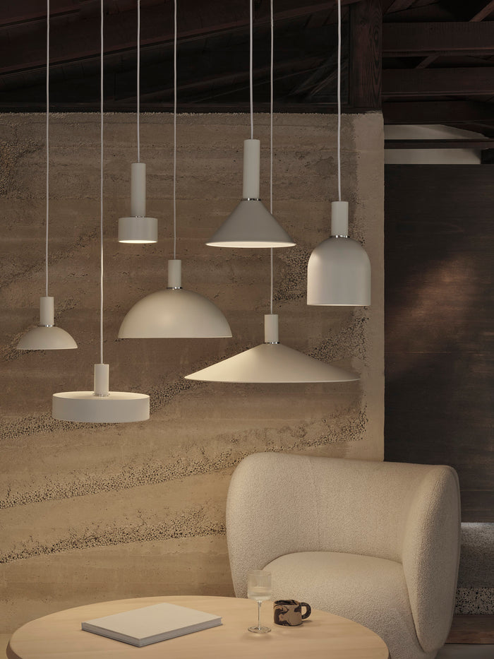 Group of pendant shades in white and light tones hung against a textured wall, with a bouclé lounge chair and round table