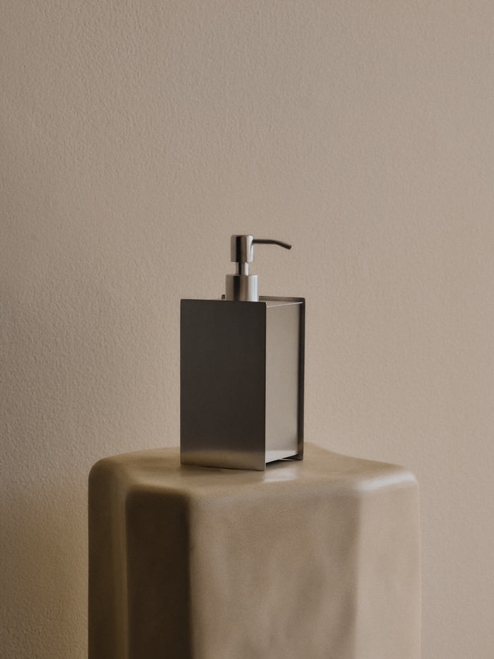 Square soap dispenser with pump standing on a rounded plinth against a beige wall