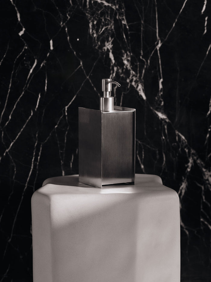 Square soap dispenser with pump on a pale plinth against a dark veined wall